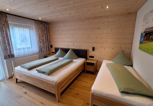 helles Schlafzimmer mit Doppelbett, Einzelbett, Holzbdoden, hellem Holz an Decke und Wand mit Fenster und Balkon