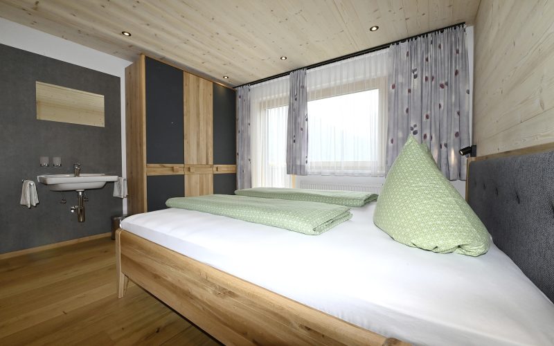 zweites Schlafzimmer mit Doppelbett, Holzboden, Heller Holzverkleidung an Wand und Decke. Dreitüriger Kleiderschrank und Waschbecken mit Spiegel.